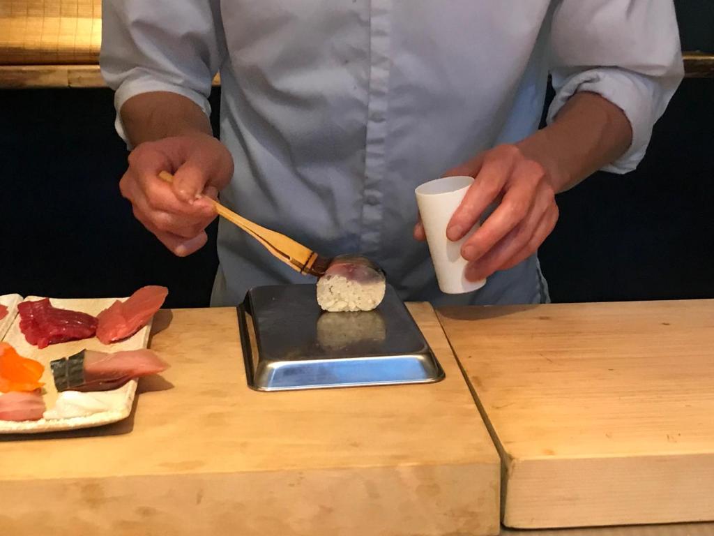 Un chef prépare des sushi avec une cuillère en bois, en tenant un morceau de poisson dans une main et une tasse dans l'autre.