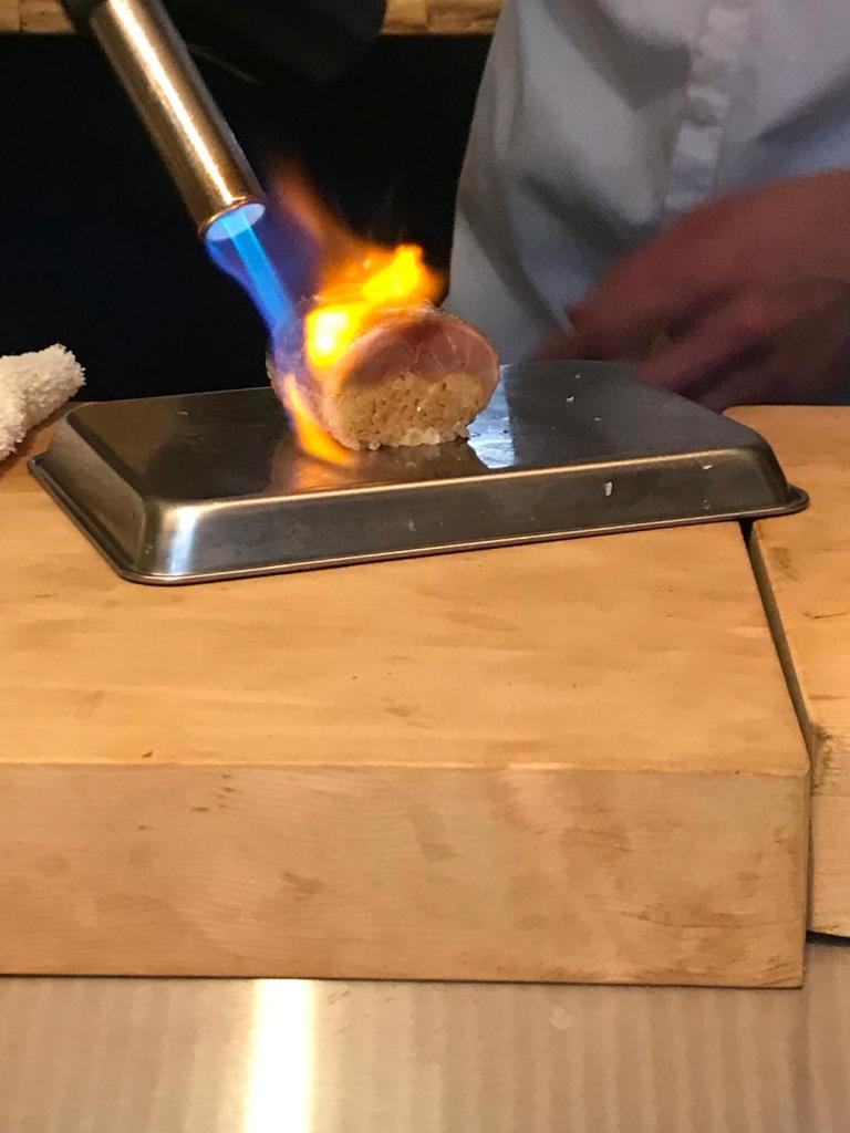 Un chef utilise un chalumeau pour flamber un morceau de viande sur une plaque en métal, à côté d'un plan de travail en bois.