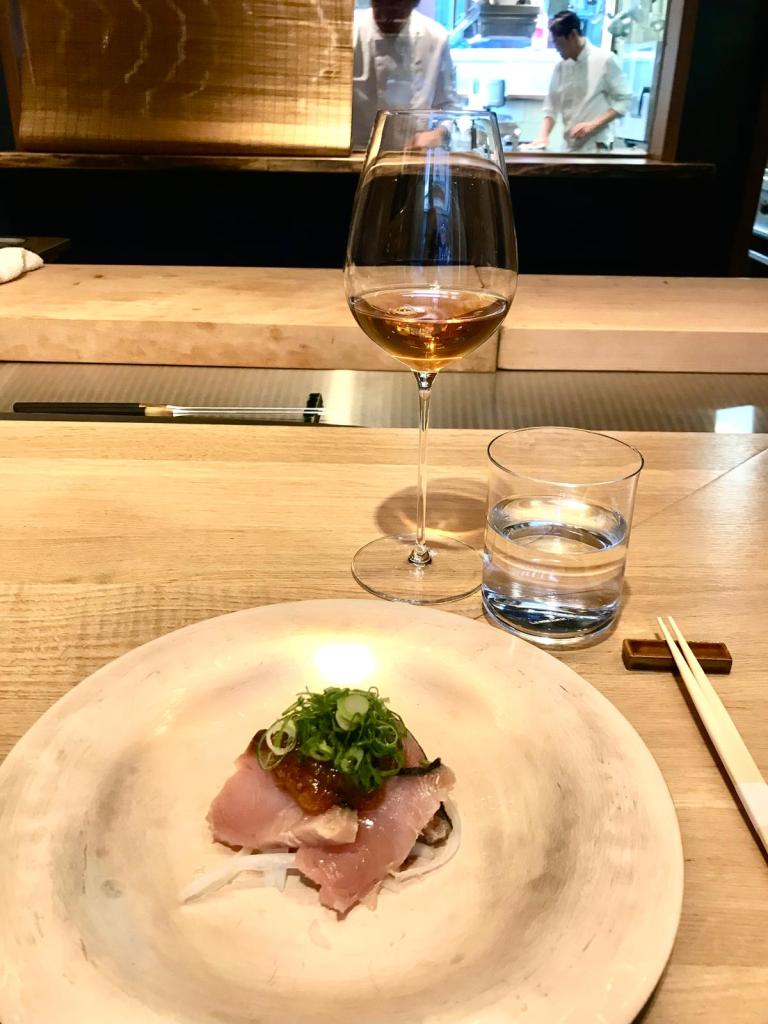 Un plat de sashimi avec des morceaux de poisson, des herbes et une sauce, accompagné d'un verre de vin blanc et d'un verre d'eau, sur une table en bois.