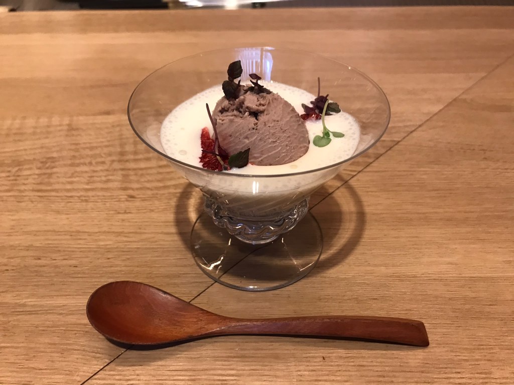 Un dessert dans un verre transparent contenant de la crème et une boule de glace au chocolat, décoré de feuilles et de fleurons, avec une cuillère en bois à côté.